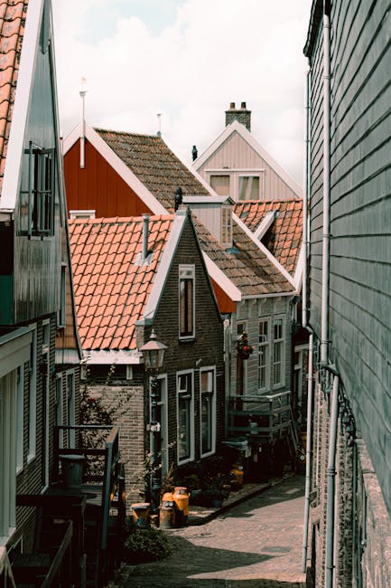 A narrow residential street with closely packed houses featuring varied architectural styles and roof designs, viewed from a balcony or elevated position. The image shows the facades and rooftops of multiple buildings, including a dark-brick house with a small front yard and a set of steps leading down to the pavement, which is strewn with cardboard boxes and plastic crates likely containing household items. Wooden and metal railings line the edges of the balconies and stairs, and a few potted plants and outdoor lamps are visible. Overhead, the sky appears overcast, casting diffused light over the scene. This setting depicts a typical home relocation or furniture transport process in a compact street, with Holland Park Removals providing moving services tailored for narrow streets, ensuring careful loading and transport of household belongings through limited space.