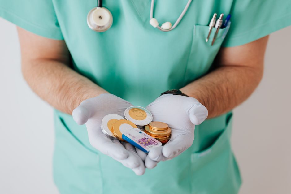 A person wearing light green scrubs with a stethoscope and pens tucked into the chest pocket is holding a transparent container filled with gold and copper coins and a few euro banknotes, which are vi