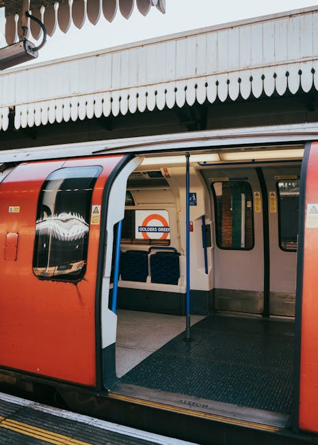 An open London Underground train carriage at a station platform, with the station name 'Golders Green' visible on the sign inside. The train interior shows dark blue upholstered seats arranged in pairs along the sides, with some seats visible close to the doors. A small step is positioned inside the entrance for ease of boarding, and the platform edge with yellow safety line can be seen outside. The exterior of the train is red with a white and black window design, and the station structure above features a white wooden canopy with decorative scalloped trim. The setting suggests a typical urban transit environment, relevant to home relocation and furniture transport in London, where Holland Park Removals might assist with moving services including train-based logistics for the W11 area.