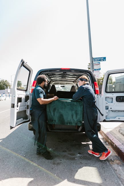 Two movers from Holland Park Removals are loading a large, rectangular piece of furniture wrapped in green fabric into the back of a white van, parked on a street near a lamppost with a blue street sign indicating Somerset Road. The van's rear doors are open, revealing the interior where the furniture is being carefully placed inside. One mover, wearing a dark uniform and safety glasses, holds the furniture steady, while the other, dressed in a blue T-shirt and black trousers, assists with the movement. The street has a paved sidewalk, and the scene is set during daylight hours with clear weather, emphasizing the process of home relocation or furniture transport as part of professional packing and moving services, with Holland Park Removals specializing in such house removals in the W11 area.