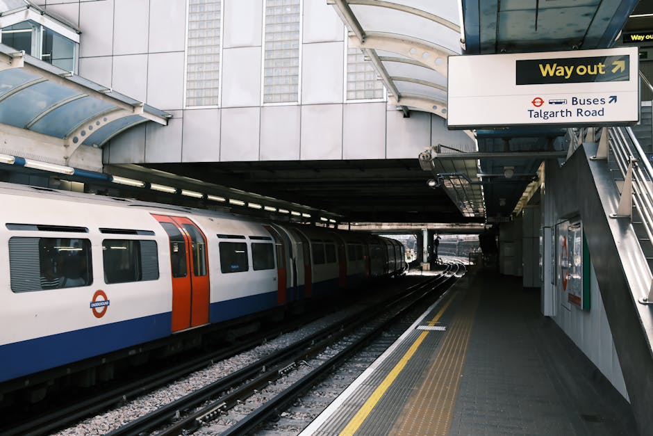 An open London Underground train carriage at a station platform, with the station name 'Golders Green' visible on the sign inside. The train interior shows dark blue upholstered seats arranged in pairs along the sides, with some seats visible close to the doors. A small step is positioned inside the entrance for ease of boarding, and the platform edge with yellow safety line can be seen outside. The exterior of the train is red with a white and black window design, and the station structure above features a white wooden canopy with decorative scalloped trim. The setting suggests a typical urban transit environment, relevant to home relocation and furniture transport in London, where Holland Park Removals might assist with moving services including train-based logistics for the W11 area.